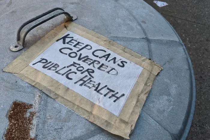 A photo of a trash can with a sign reading, "keep cans covered for public health"